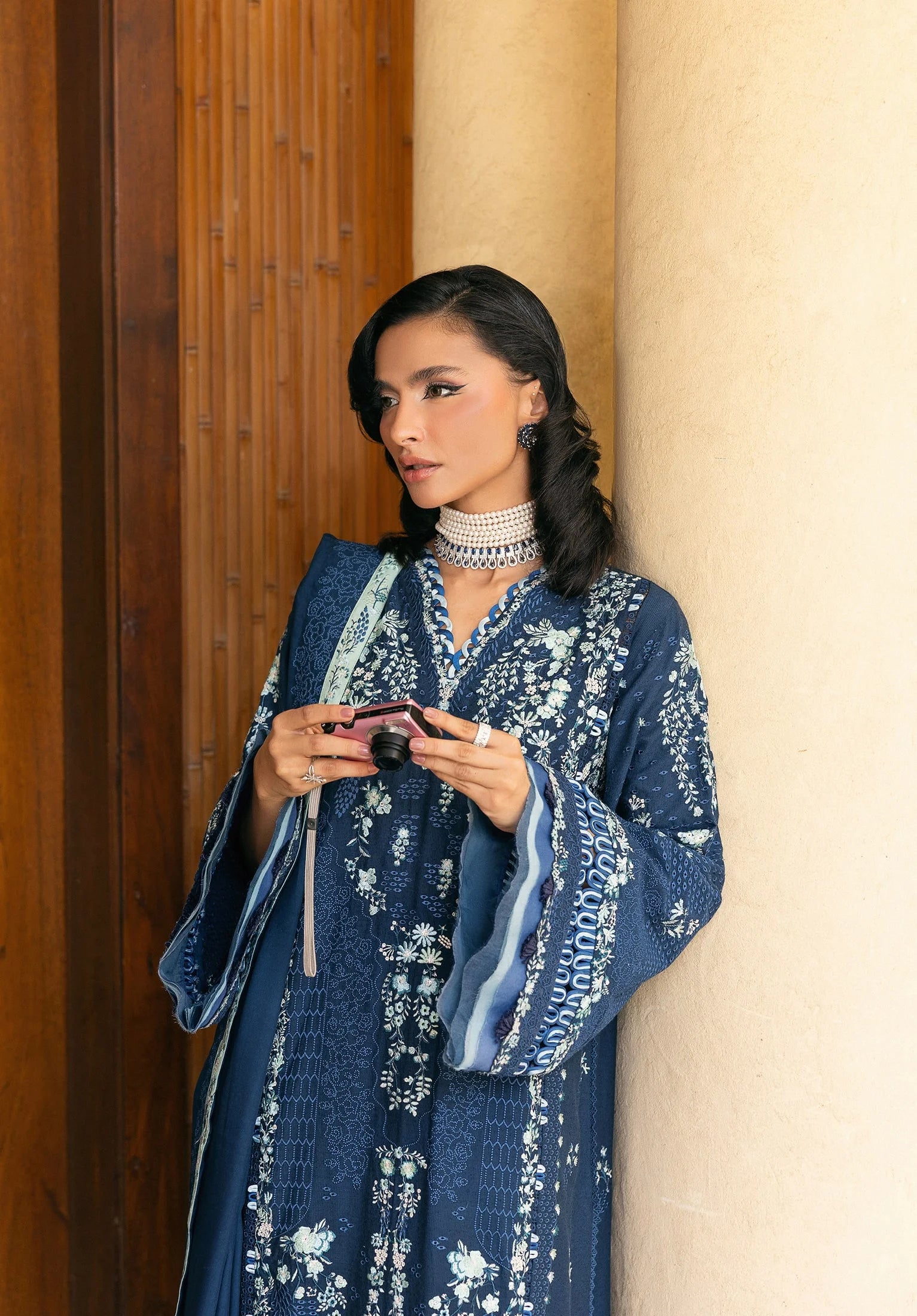 Woman in a blue embroidered outfit standing against a beige wall.