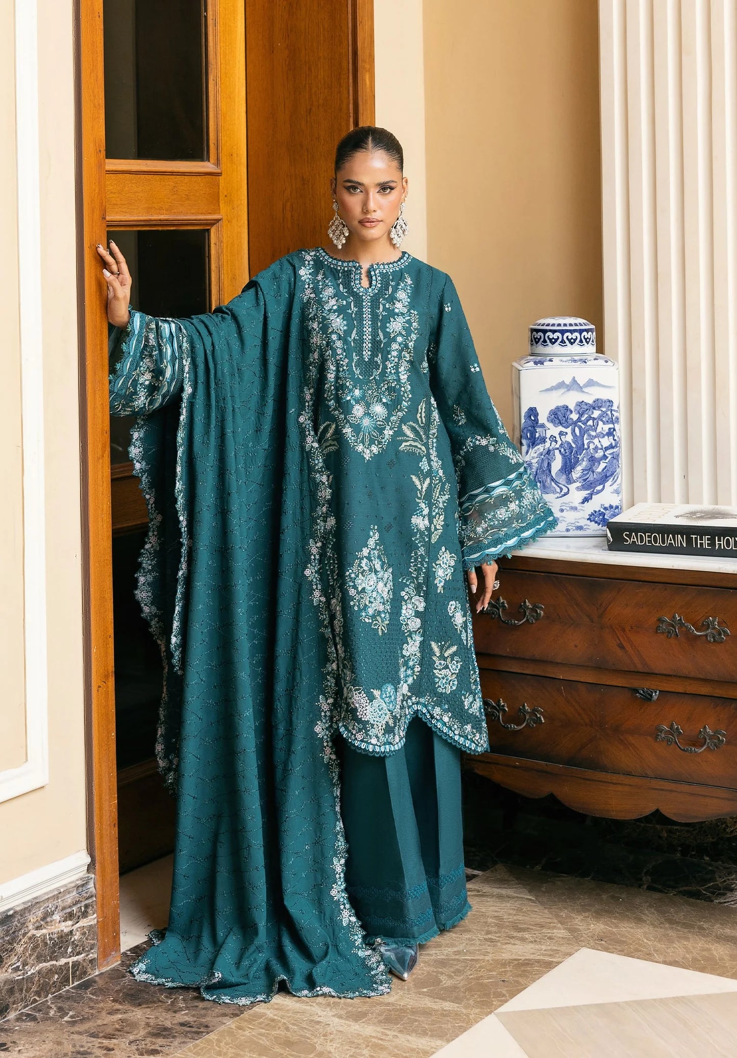 Woman wearing a teal embroidered dress standing in a room with wooden furniture and decorative items from LUXURY WINTER '25