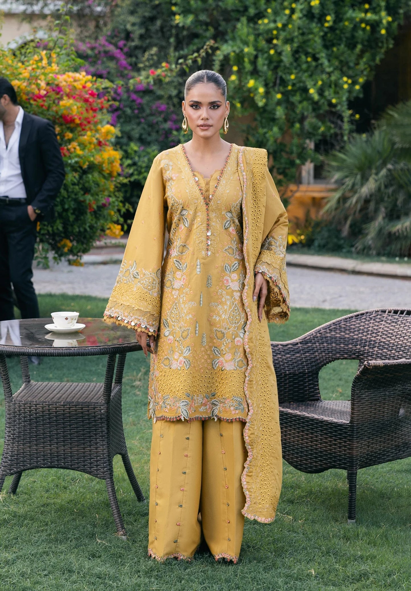 Woman in a yellow embroidered outfit standing outdoors with greenery and furniture in the background