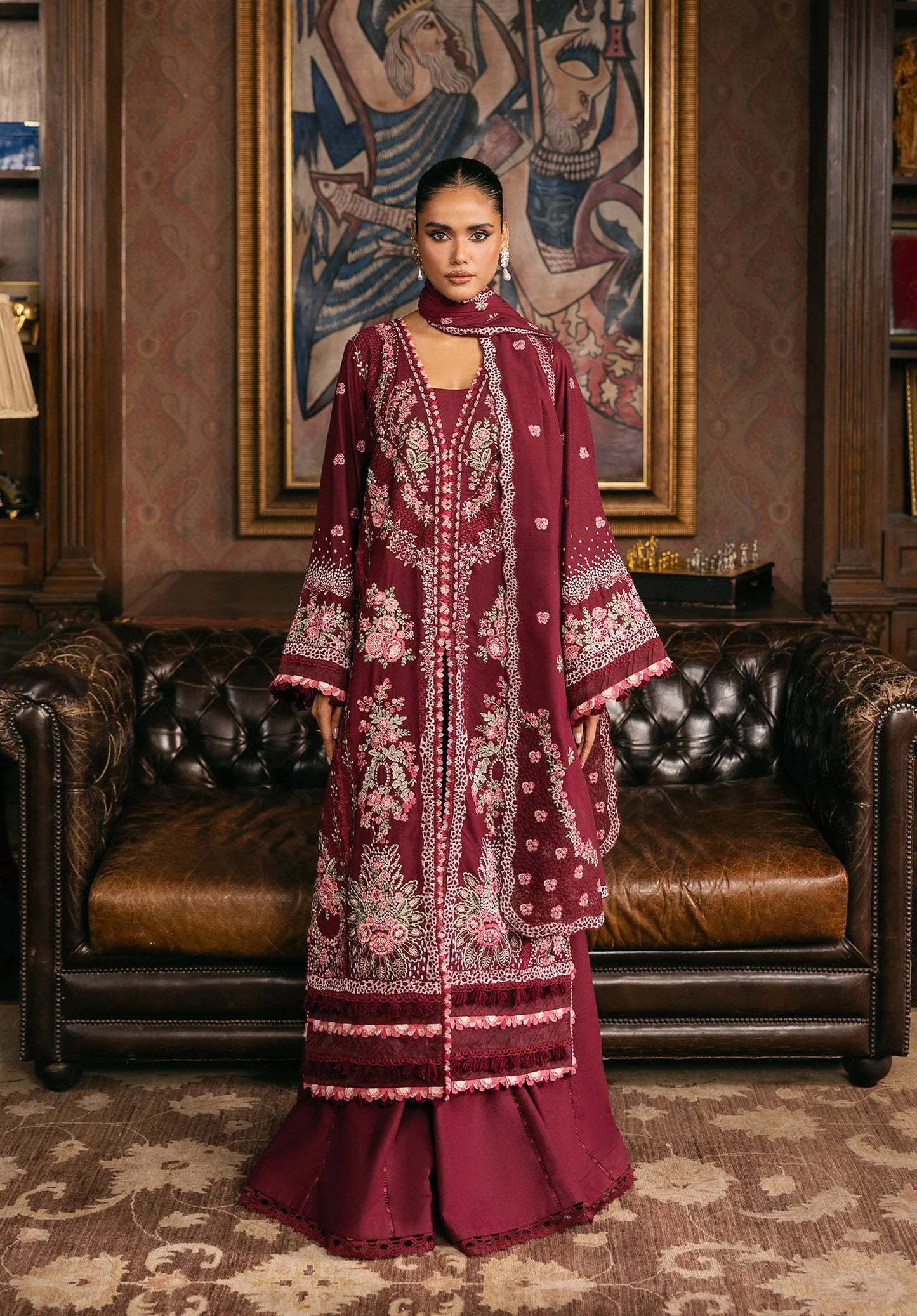 Woman in a burgundy embroidered dress standing in an elegant room with leather furniture and a decorative wall.