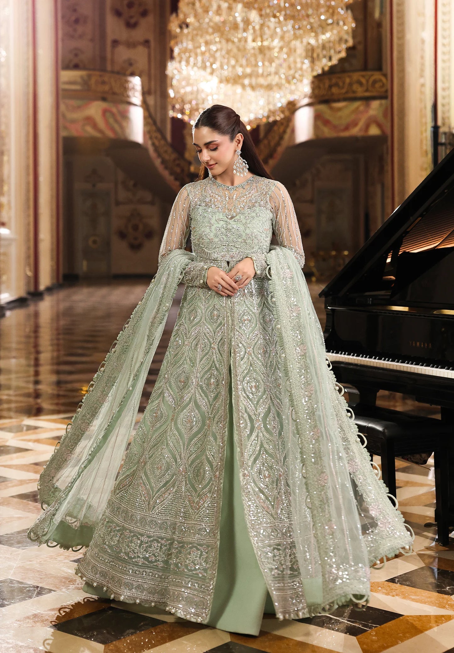 Woman in a light green embroidered traditional outfit standing in an elegant interior setting with a chandelier and piano.
