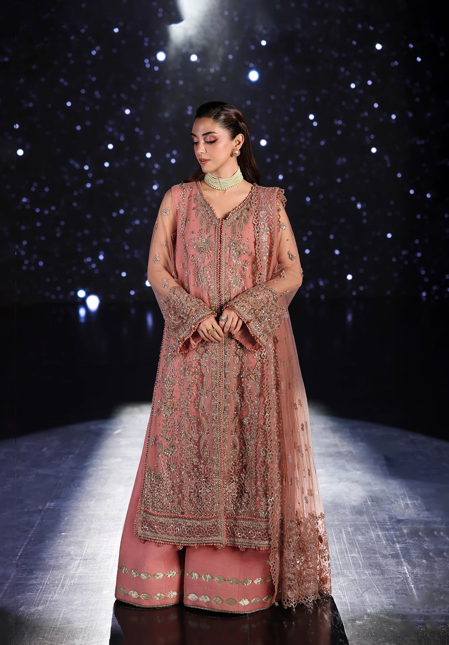 Woman in a traditional pink embroidered outfit on a dark background with star-like lights.