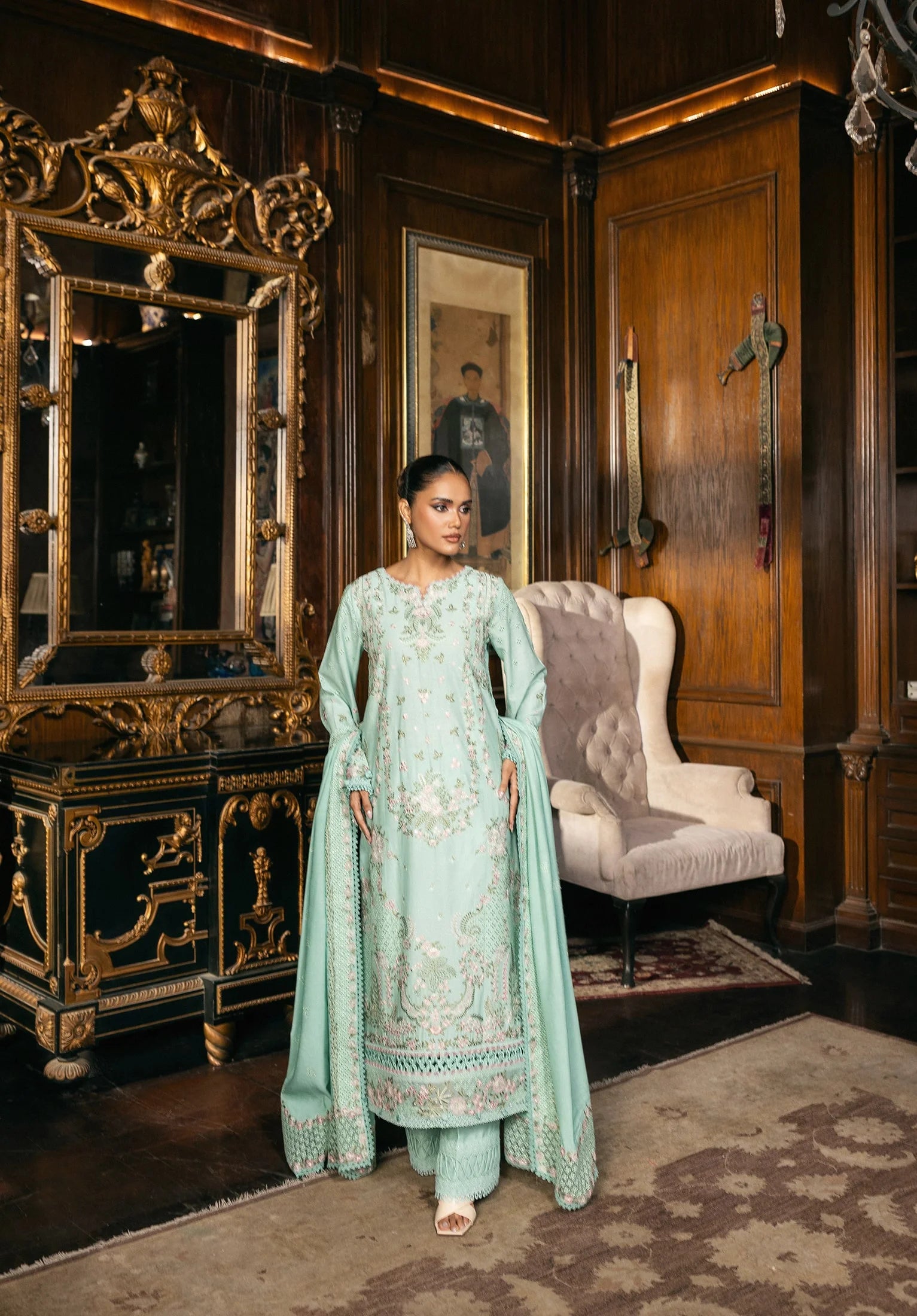 Woman in a light blue traditional outfit standing in an ornate room with wooden furniture and a large mirror.