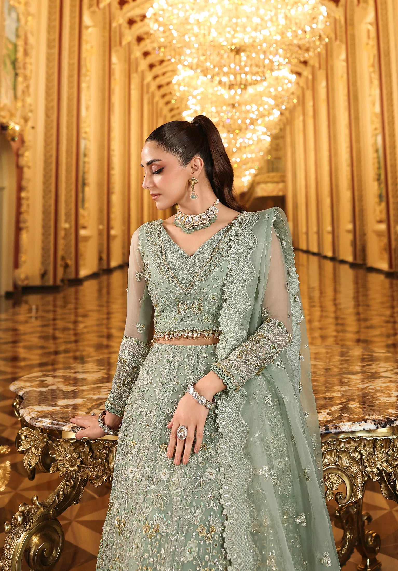 Woman in a light green embroidered traditional outfit in an ornate room with chandeliers.