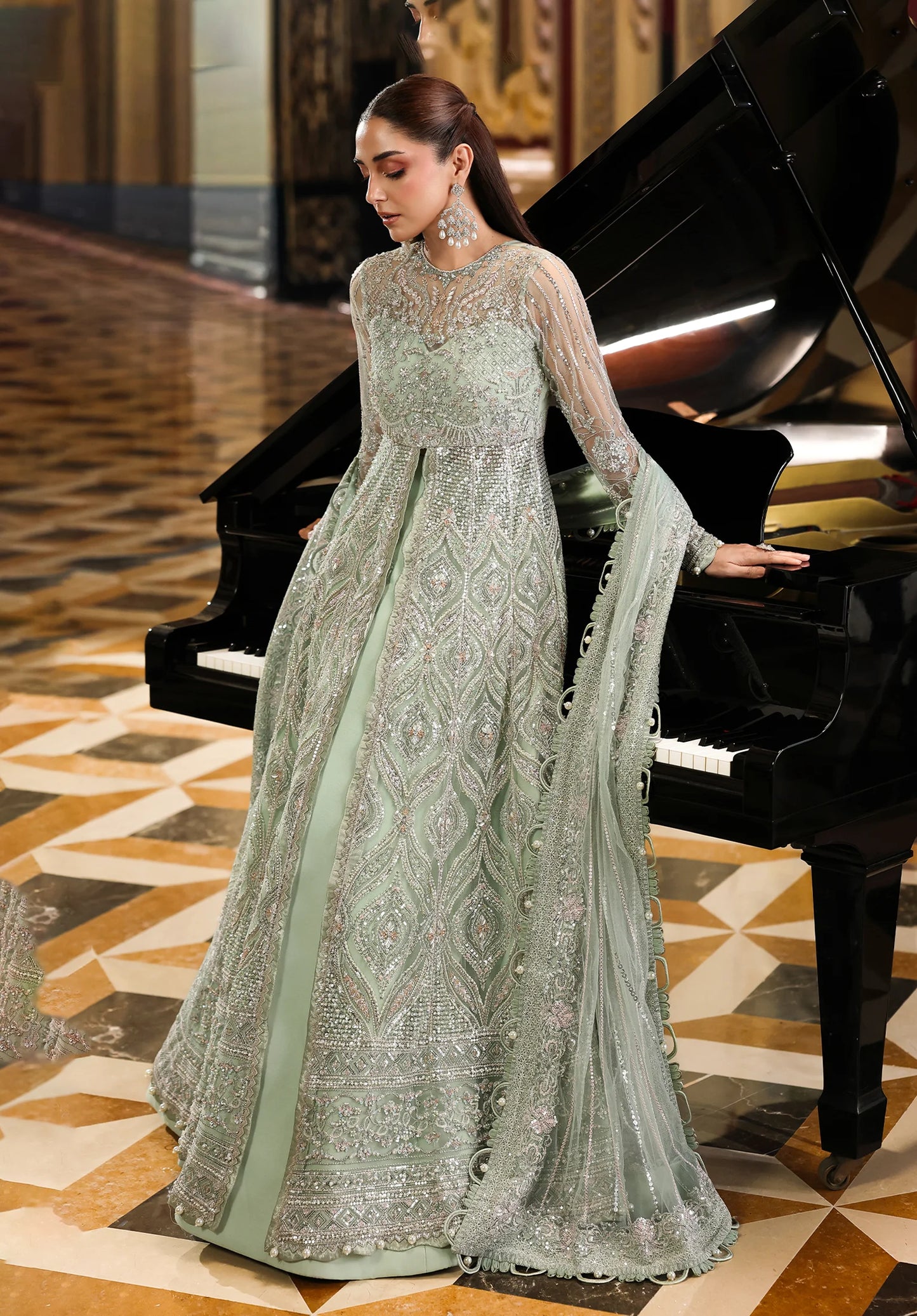 Woman in a light green embroidered dress standing next to a grand piano in an elegant interior setting.