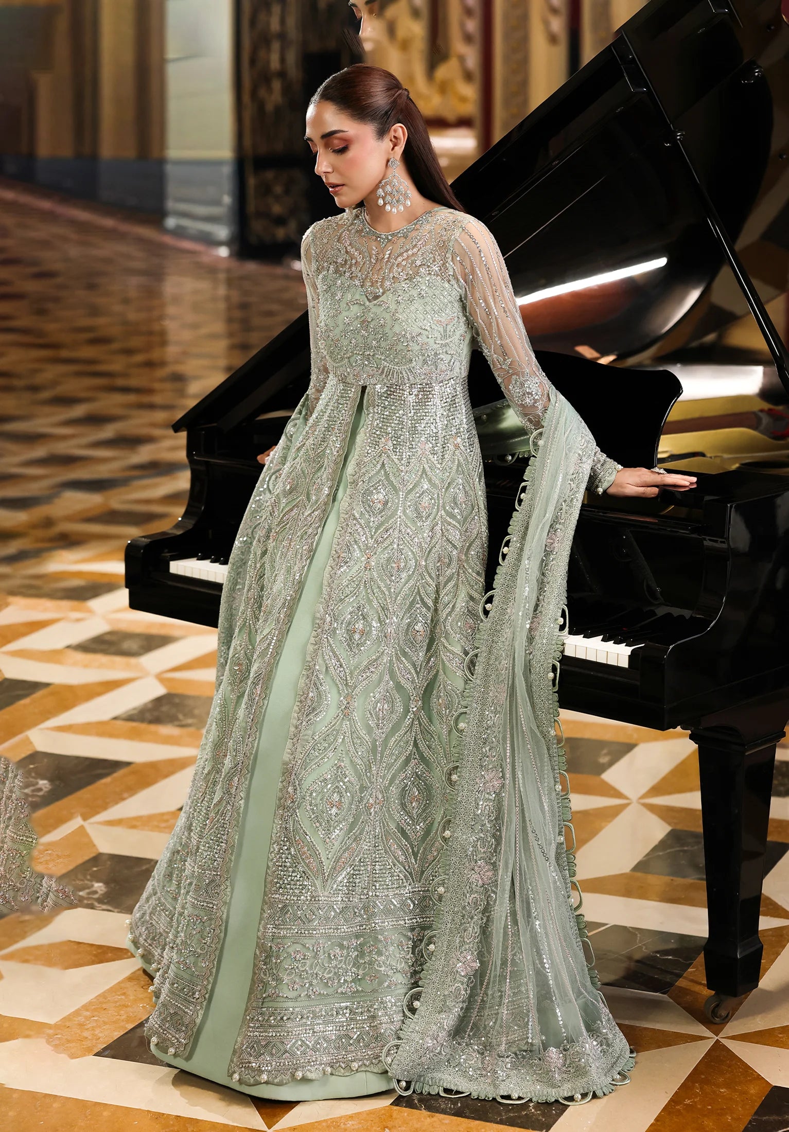 Woman in a light green embroidered dress standing next to a grand piano in an elegant interior setting.